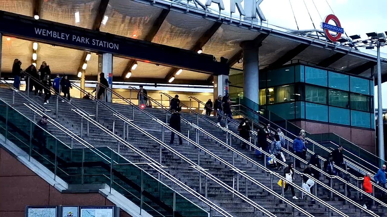 People walking down the broad outdoor staircase at Wembley Park station, with the station sign and an Underground roundel on the canopy