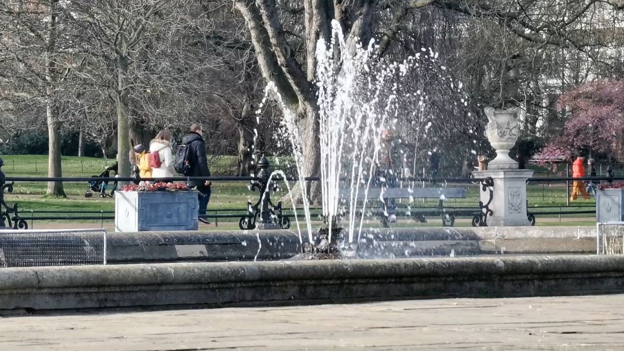 A fountain in Hyde Park with bare trees behind and people walking past