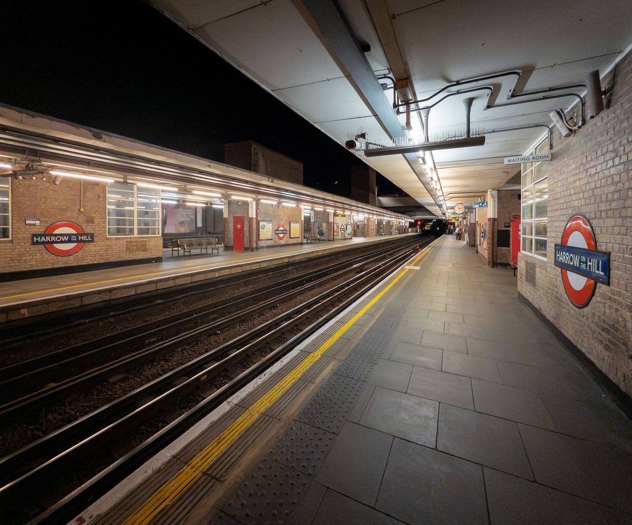 Harrow-on-the-Hill roundel on a brick wall beside an empty platform at night, with a train at the far platform