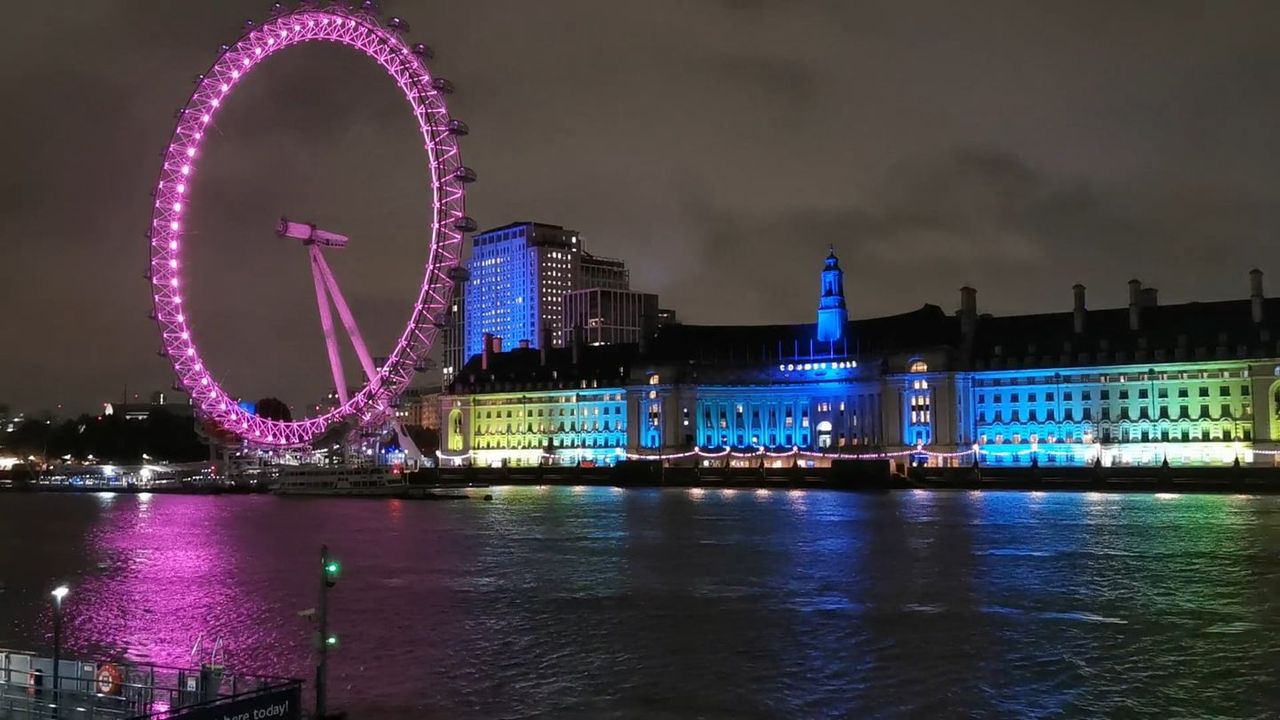 The London Eye lit pink at night, seen across the Thames with lit buildings along the far bank